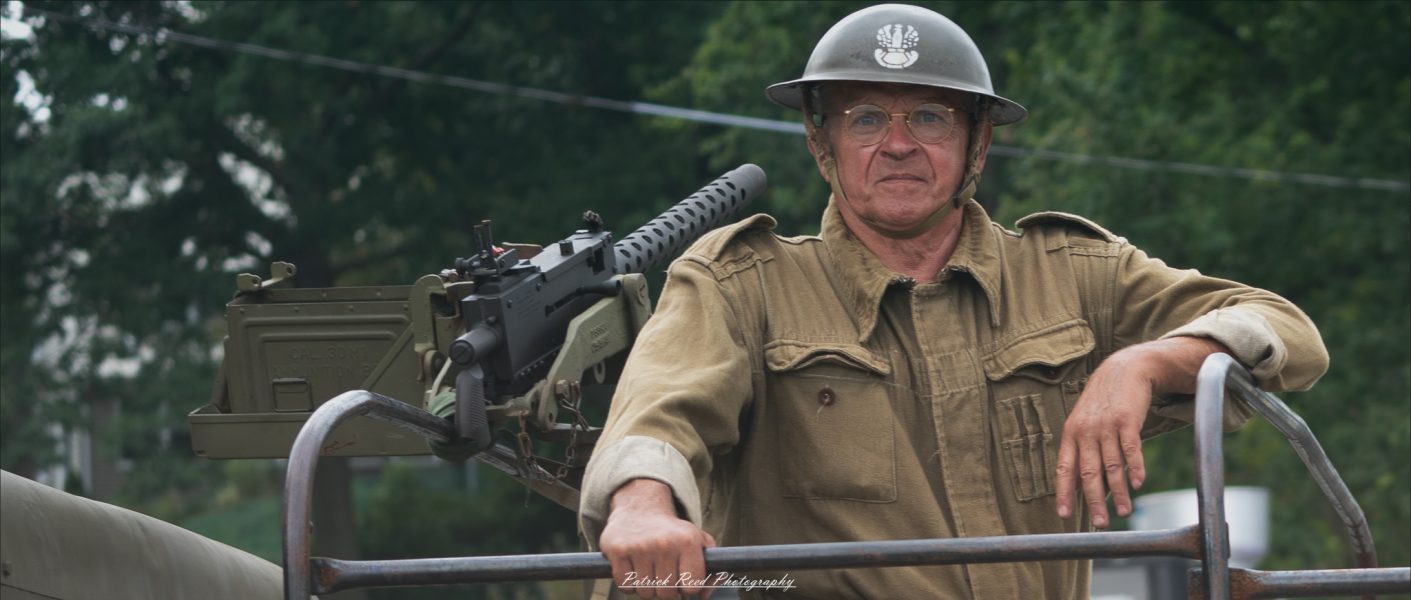A soldier on a truck, with a .50 caliber machine gun mounted in the background. The image reflects readiness and strength, highlighting the soldier’s role in a heavily armed convoy, prepared for action.